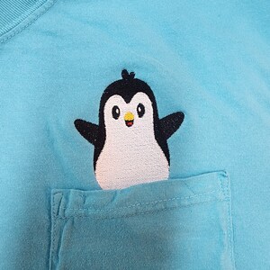 May include: A light blue t-shirt with a small embroidered penguin on the front pocket. The penguin is black and white with a yellow beak and a happy expression.