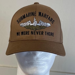 May include: A brown baseball cap with the text "SUBMARINE WARFARE" and "WE WERE NEVER THERE" embroidered in black. The cap features a white embroidered submarine emblem.