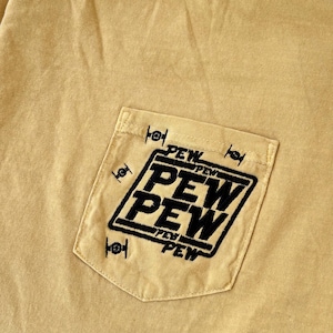 May include: Mustard yellow t-shirt with a pocket featuring the embroidered text "PEW PEW" in a black, bold font. The pocket design includes small black starfighter graphics. The shirt is made of a soft, casual fabric.