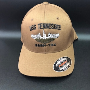 May include: A tan Flexfit baseball cap with the text "USS TENNESSEE" embroidered on the front, along with an emblem and the number "SSBN-734". The cap has a curved brim and a Flexfit sticker.