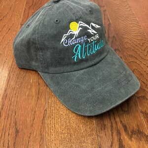 K&ouml;nnte beinhalten: Eine dunkelgraue Baseballkappe mit einem gestickten Design aus Bergen, einer Sonne und dem Text "Change Your Altitude".