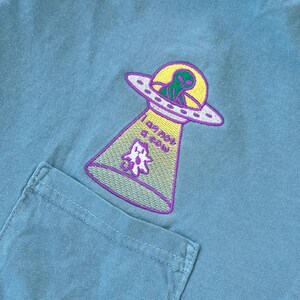 May include: A light blue t-shirt with an embroidered alien in a UFO. The UFO is purple and yellow with the text "I am not a clown" written on the side.