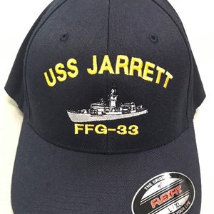 May include: A navy blue baseball cap with yellow embroidered text that reads "USS JARRETT FFG-33". A white embroidered image of a ship is above the text. The cap has a black Flexfit label on the side.
