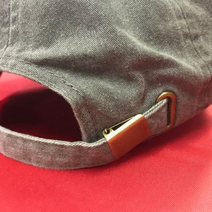 K&ouml;nnte beinhalten: Eine graue Baseballkappe aus Baumwolle mit einem verstellbaren Riemen aus Messing und einer Schnalle.