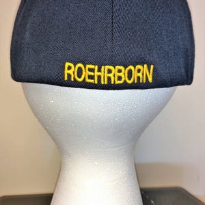 May include: Navy blue baseball cap with the word "ROEHRBORN" embroidered in yellow thread across the back. The cap is displayed on a white mannequin head.