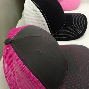 May include: Three trucker hats with mesh back panels. The top hat is pink with black mesh. The middle hat is black with white mesh. The bottom hat is gray with pink mesh.