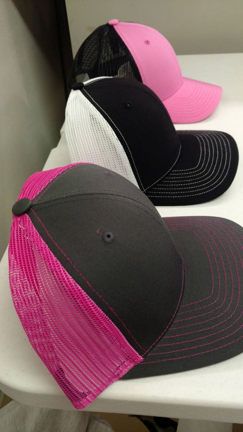 May include: Three trucker hats with mesh back panels. The top hat is pink with black mesh. The middle hat is black with white mesh. The bottom hat is gray with pink mesh.