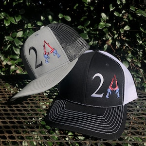 May include: Two trucker hats with a mesh back. One hat is gray with black mesh and the other is black with white mesh. Both hats have the number "2A" embroidered on the front with a red, white, and blue American flag design.