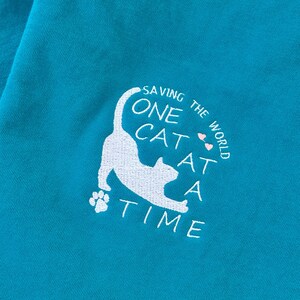 May include: A turquoise blue t-shirt with a white embroidered design of a cat with the text "Saving the World One Cat at a Time".