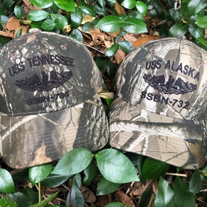 May include: Two camouflage baseball caps with black embroidered text and images. The caps say "USS Tennessee SSBN-764" and "USS Alaska SSBN-732".