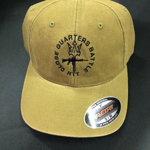 May include: A tan baseball cap with a black embroidered logo that says "Close Quarters Battle" with a trident and rifle design. The cap has a black Flexfit label on the side that says "The Original Flexfit S/M It Fits Your Life."