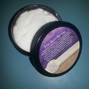 Peut inclure: Un pot noir avec un couvercle blanc contenant 237 ml de beurre corporel. Le pot est doté d'une étiquette violette avec du texte blanc qui indique "8 oz. Body Butter" et liste les ingrédients. L'étiquette comprend également le nom "3 Gypsy's Spa Apothecary LLC" et une adresse de site Web.