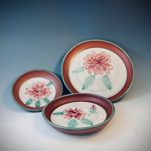 Pode incluir: Três pratos de cerâmica com fundo branco e design de flores vermelhas. Os pratos estão aninhados um dentro do outro, com o prato maior na parte inferior e o menor na parte superior.