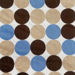 May include: A close-up of a fabric with a repeating pattern of large circles. The circles are in shades of brown, tan, and blue, set against a white background. The design creates a playful and modern aesthetic, suitable for various textile applications.