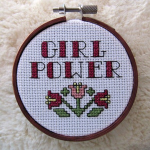May include: A white embroidery hoop with a red and black cross-stitch design that reads "GIRL POWER" with three red flowers and green leaves below the text.