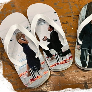 Puede incluir: Un par de chanclas blancas con fotos personalizadas. Las chanclas muestran una pareja en un entorno de boda. La chancla izquierda muestra a la mujer con un vestido negro, mientras que la chancla derecha muestra al hombre con un traje y a la mujer con un vestido blanco. Las chanclas tienen las iniciales "TH" impresas.