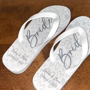Könnte beinhalten: Weiße Flip-Flops mit einem Herzmuster und dem Wort "Bride" in Grau. Die Flip-Flops sind mit den Namen "Debra & Steve" und dem Datum "October 5, 2023" personalisiert.