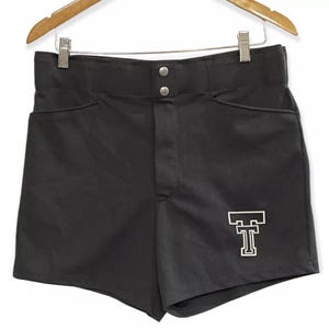 May include: Black athletic shorts with a white embroidered 'T' on the right leg. The shorts have a button fly and two pockets.