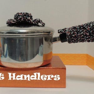 May include: A stainless steel saucepan with a lid, featuring a black, white, and red crocheted handle cover and a matching crocheted knob cover on the lid. The base of the pot rests on a box with the text Pot Handlers printed in white.