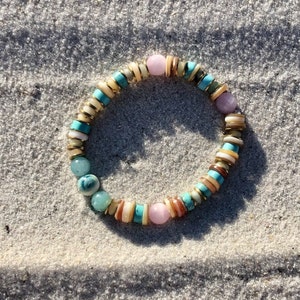May include: A colorful beaded bracelet with turquoise, pink, and gold beads. The bracelet is on a white sandy surface.