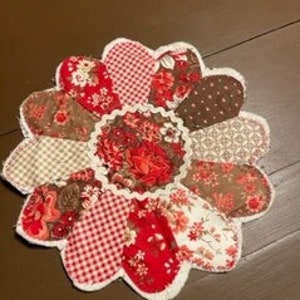 Op de afbeelding: Een rode, bruine en witte patchwork-onderzetter in de vorm van een bloem met een gegolfde rand.