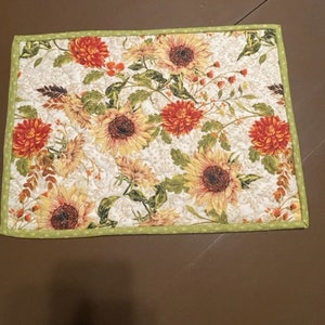 Könnte beinhalten: Ein grünes und weißes gestepptes Platzset mit einem Blumenmuster aus Sonnenblumen und roten Blumen. Das Platzset hat einen grünen Rand mit weißen Punkten.