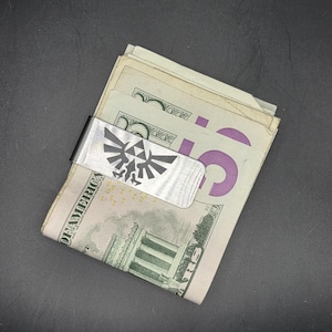 May include: A silver money clip with a black and white design of a Triforce symbol, holding several U.S. dollar bills. The clip is made of brushed metal and is set against a dark background.
