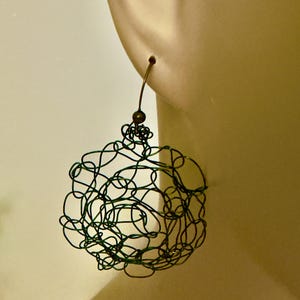 May include: A spherical earring constructed from thin, dark green metallic wire formed into a chaotic, tangled web pattern. The wire sphere hangs from a simple, curved bronze-toned metal hook that passes through the top of the earring structure.