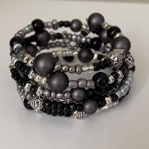 May include: A multi-strand bracelet with a wrap-around design. The bracelet features a mix of black, gray, and silver beads. Some beads are matte gray spheres, while others are small, shiny silver or black. Decorative silver metal beads are also included.