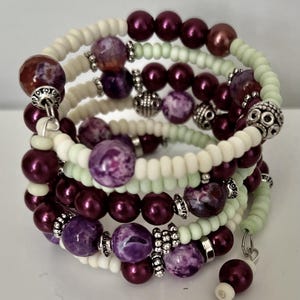 May include: A multi-strand wrap bracelet featuring a mix of beads. The bracelet includes deep purple, white, and light green beads. Accents include silver-colored metal beads and a dangling charm. The bracelet is designed to wrap around the wrist multiple times.