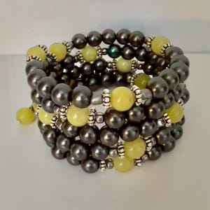 May include: A multi-strand bracelet featuring dark gray pearls and light yellow beads. The bracelet is accented with silver-colored metal spacers. The design is a wrap-around style, creating a layered effect. The beads are round and evenly spaced.