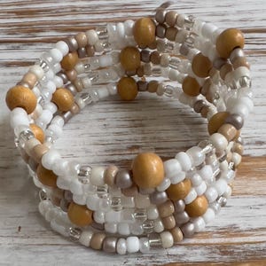May include: A multi-strand wrap bracelet featuring white, tan, and brown beads, interspersed with wooden beads. The bracelet is designed to wrap around the wrist multiple times. The beads are strung together to create a textured, layered effect.