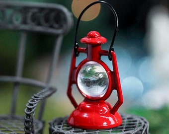 May include: A miniature red lantern with a clear glass globe and a black handle. The lantern is sitting on a black metal table.