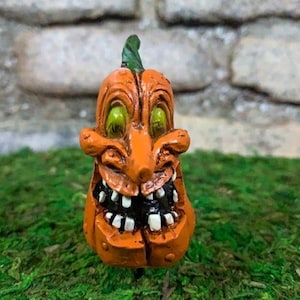 May include: A small orange pumpkin figurine with a green stem and a carved, toothy grin. The pumpkin has green eyes and a crooked nose.