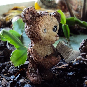 May include: A brown teddy bear figurine holding a small wooden axe. The bear has black eyes and a brown fur texture. The figurine is sitting on a bed of brown mulch.