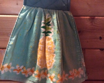 May include: A teal kitchen towel with a pineapple design and a white button on the top. The towel has a white and yellow floral border.