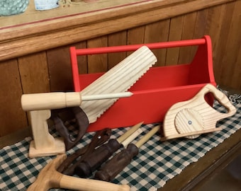 May include: A red wooden toolbox filled with wooden toy tools, including a saw, hammer, and drill. The tools are arranged on a green and white checkered cloth. The background features a floral wallpaper.