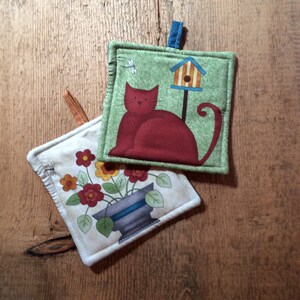 May include: Two fabric pot holders. One is green with a red cat and a birdhouse. The other is white with a flower pot and flowers.
