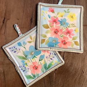 Könnte beinhalten: Zwei Topflappen mit floralem Muster auf hellblauem Hintergrund und hellbraunem Rand. Die Topflappen haben einen Schlaufe und zeigen ein Aquarell-Blumenmuster mit rosa, blauen und gelben Blumen.