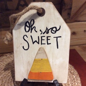 May include: A white wooden sign with a rope handle and the text "Oh, so SWEET" in black paint. A painted candy corn design is below the text.