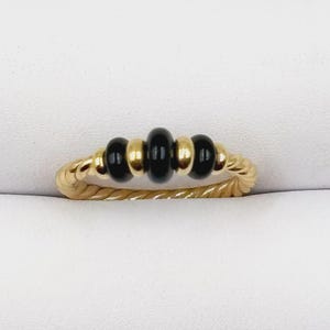 May include: A gold ring with a twisted band design. The ring features three black beads and two gold beads in the center. The ring is set against a white background.