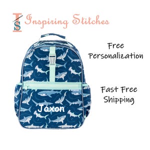 May include: A dark blue backpack featuring a repeating pattern of various sharks in light blue and white. The bag includes a light blue vertical strap with a plastic buckle, a front zippered pocket, and the name Jaxon embroidered in white.