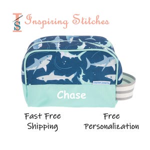 May include: A blue and teal shark-themed travel bag with the name "Chase" printed on the front. The bag features a shark pattern on the top portion and a teal bottom. It has a gray and white striped handle and the text "Inspiring Stitches".