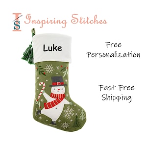 Peut inclure: Chaussette de Noël verte avec un revers blanc et un motif de bonhomme de neige. Le bonhomme de neige porte une écharpe rouge et un chapeau noir, tenant un sucre d'orge. Le nom "Luke" est brodé sur le revers. La chaussette est décorée de flocons de neige et d'arbres.
