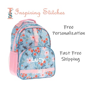 May include: A light blue backpack with a floral and bee pattern. The backpack has pink accents on the straps, handle, and side pocket. The name "Lucy" is printed on the front. The text "Inspiring Stitches" is at the top.