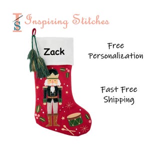 May include: A red Christmas stocking with a white cuff, featuring a nutcracker design. The stocking has the name "Zack" printed on it, along with green tassels and a drum and stars design. The text "Inspiring Stitches" is also visible.