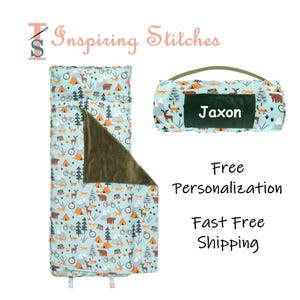 May include: A light blue sleeping bag and matching carrying case with a camping-themed pattern featuring tents, bears, and bicycles. The carrying case has a green handle and a personalized name tag that says "Jaxon".