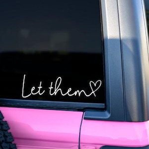 May include: A pink car with a black window and a white vinyl decal that says "Let them" with a heart symbol.