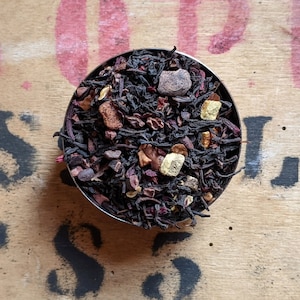 May include: A close-up of a metal bowl filled with a blend of black tea, chocolate pieces, dried fruit, and other ingredients. The tea blend is a dark brown color and the ingredients are scattered throughout the bowl.