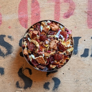 Op de afbeelding: Een close-up van een kom gevuld met een mengsel van gedroogd fruit, bloemen en kruiden. Het mengsel bevat sinaasappelschil, appelstukjes, cranberries en rozenblaadjes.